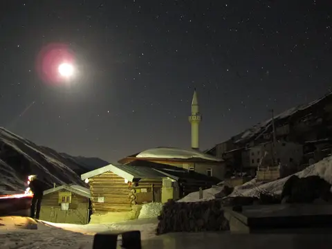 Kar kaplı dağ köyünde gece aydınlatılmış minareli cami ve çevresindeki evler
