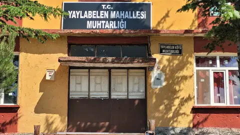 Yaylabelen mahalle muhtarlığı tabelası asılı, sarı ve kırmızı boyalı iki katlı binanın cephesi