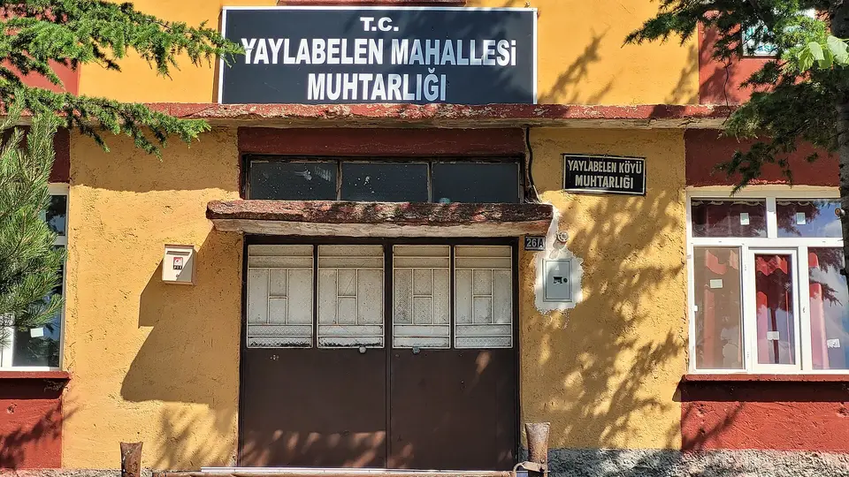 Yaylabelen mahalle muhtarlığı tabelası asılı, sarı ve kırmızı boyalı iki katlı binanın cephesi