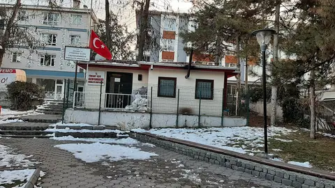 Karla kaplı zeminde bayrak direği olan tek katlı mahalle muhtarlığı binası ve arkasındaki apartmanlar