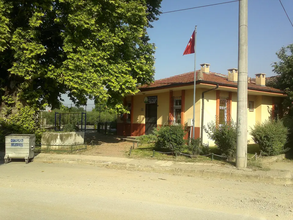 Çeltik'te ağaçların gölgesinde tek katlı mahalle muhtarlığı binası ve bayrak direği